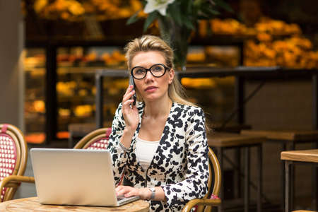 Attractive blonde woman sitting in outdoor cafe with laptop talking on the mobile phone.の写真素材