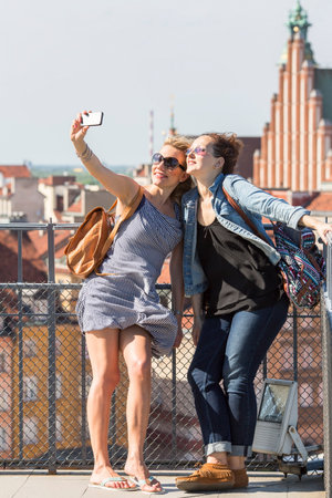 Two young female friends taking selfies on a smartphone on the background of the Castle Square in Warsaw, Poland.のeditorial素材