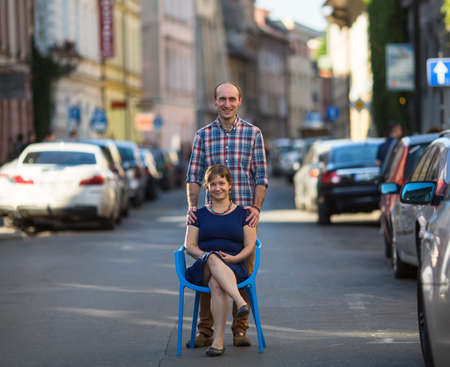 Couple, happy young family. Conceptual photo, a young woman sits on a chair in the middle of the street, a man stands behind her. European city blur in the background.の写真素材