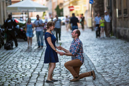 Couple in love on the street. Man on his knees gives a woman a flower, makes an offer. Man and woman during honeymoon.の写真素材