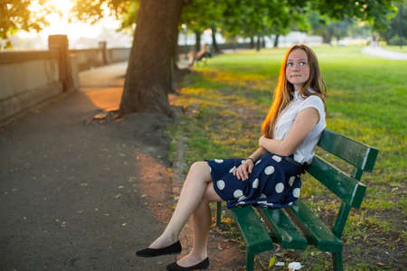 Cute young girl with long golden hair sitting alone on a bench in the Park.の写真素材