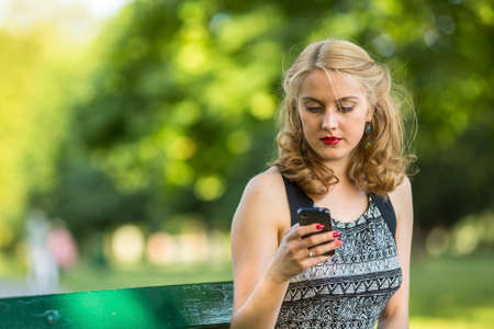 Beautiful young woman typing on smartphone, sitting on bench outdoors.
