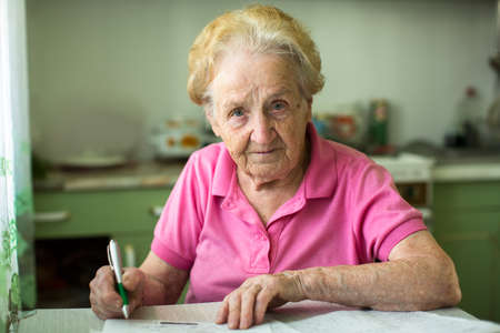 Elderly senior woman populates handle her utility bills notices, sitting at the table in the kitchen.の写真素材