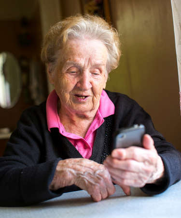 Old woman takes on smartphone sitting at the table in the house.