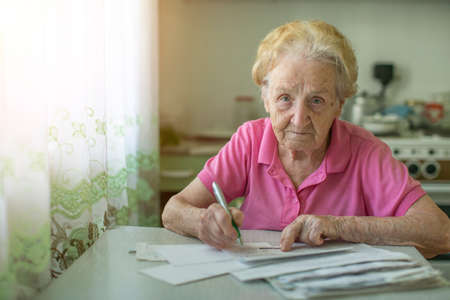 An elderly woman fills out a receipt for payment of utilities, sitting in the kitchen in house.の写真素材