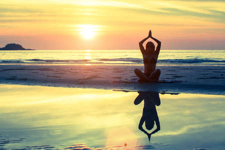 Silhouette of girl in yoga pose sitting on the beach during sunset.の写真素材