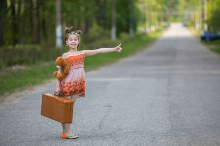 Funny little girl hitchhiker with a suitcase and a teddy bear.の写真素材
