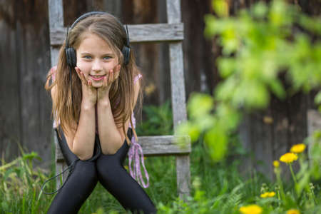 Cute girlie enjoys music with headphones on sitting in the courtyard of a village house.の写真素材