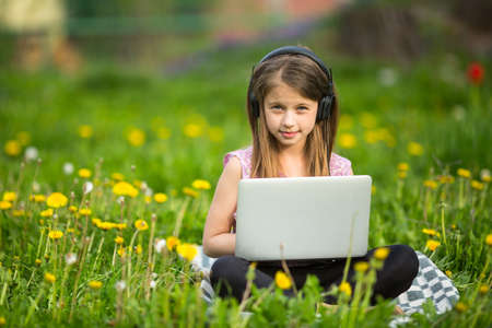 Little girl in headphones with laptop sitting on the grass in the Park.