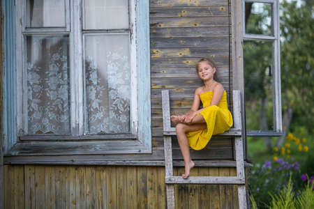 Little cute girl sitting on a wooden ladder near a village house.の写真素材