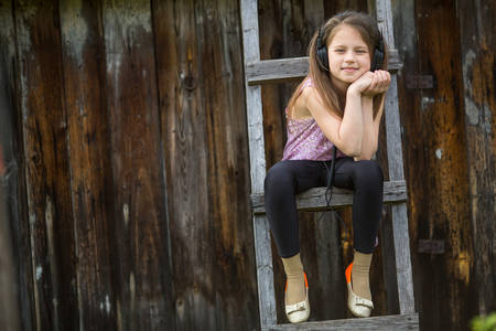 Little naughty girl with headphones sitting on a wooden stepladder in the village outdoors.の写真素材