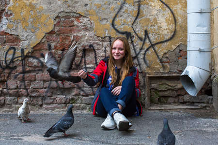 Teen girl sits near an old brick wall and feeding pigeons.の写真素材