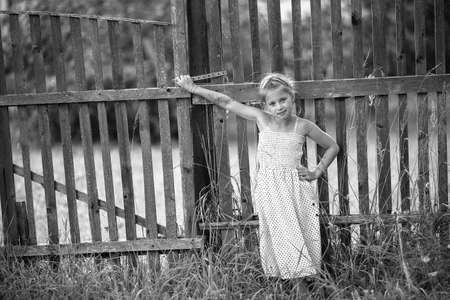 Cute little girl stands near a fence in the village. Black and white photo.の写真素材