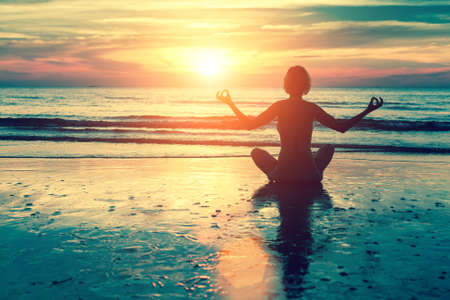 Silhouette young woman practicing yoga on the beach at ocean sunset.の写真素材