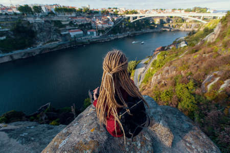 Girl with blonde dreadlocks sitting on a rock and looks at Douro river, Porto, Portugal.の写真素材
