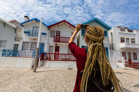 Striped houses in Costa Nova, Portugal. Young woman with blond dreadlocks makes a photo on a...