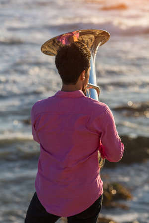 View from the back young musician play the trumpet on rocky sea coast during surf. Tuba instrument.の写真素材