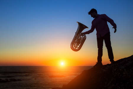 Silhouette of musician with the trumpet on rocky sea coast during amazing sunset. Tuba instrument.の写真素材