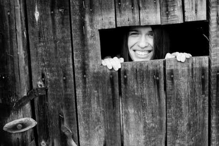 Young attractive positive woman peeking from the crack in the wooden shed.