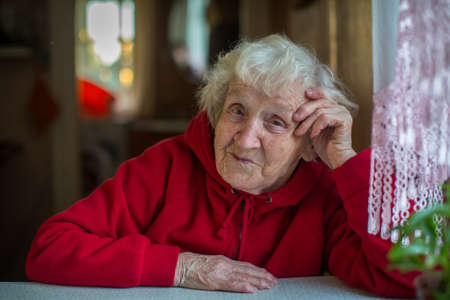 A gray-haired elderly woman in red jacket sitting at the table.の写真素材