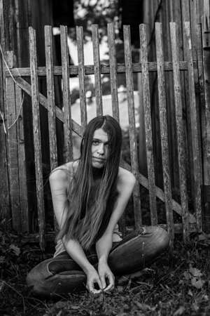Young woman with long hair in jeans sitting near the wooden fence. Black-and-white photo.の写真素材