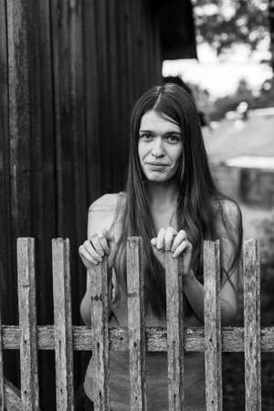 Beautiful long haired girl near the wooden fence in the village. Black-and-white photo.の写真素材