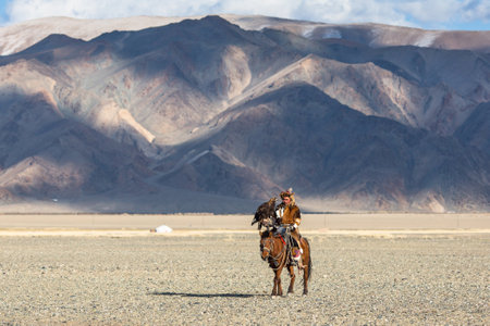 BAYAN-ULGIY, MONGOLIA - SEP 27, 2017: Kazakh Eagle Hunter at traditional clothing, on horseback while hunting to the hare holding a golden eagle on his arm in desert mountain of Western Mongolia.のeditorial素材
