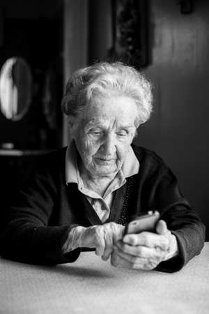 Old woman takes on smartphone sitting at the table in the house. Black-and-white photo.の写真素材