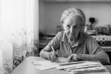 Elderly woman fills out utility bills sitting in the kitchen. Black-and-white photo.の写真素材