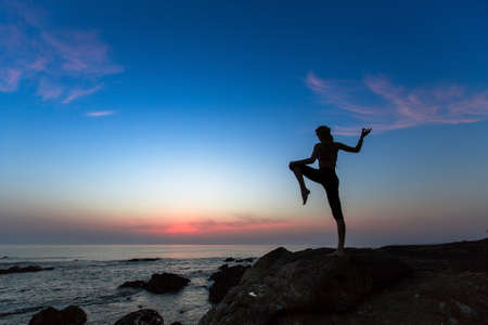 Yoga silhouette of a young woman practicing exercises on the ocean at sunset.の写真素材