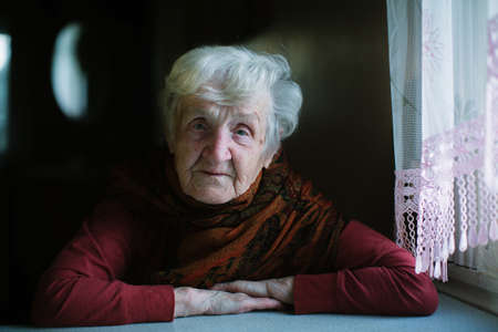 Portrait of an elderly woman in a dark key sitting in her house near window.