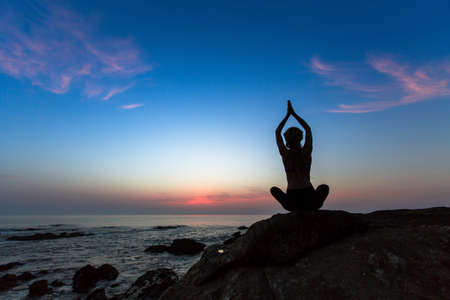 Silhouette of a young woman practicing yoga exercises on the ocean at twilight.