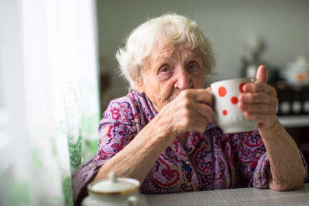 An elderly woman drinks tea sitting in the kitchen.の写真素材