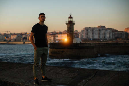 Young man near the sea lighthouse on the promenade at sunset time.