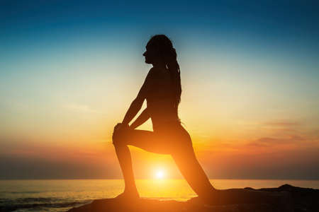 Silhouette of a young woman practicing yoga exercises on the ocean at amazing sunset.