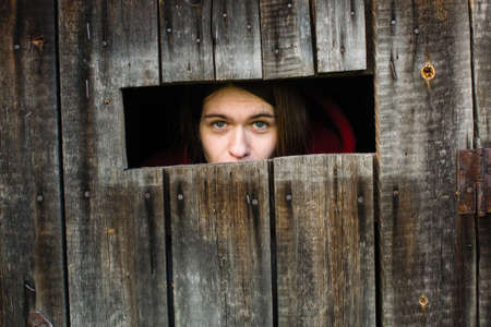 Young woman locked in a wooden old barn, sad looks out the slit.