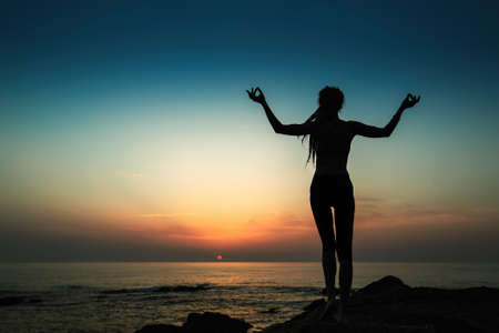 Silhouette of a flexible yoga woman stands on the shore of the evening ocean.の写真素材