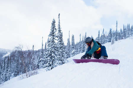 Female snowboarder buttons fastening a snowboard sitting on a snowy slope.の写真素材