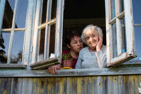 An elderly woman and her adult daughter looking out the window of the village house.の写真素材