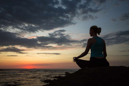 Silhouette of a woman doing yoga in Lotus pose on the twilight sea coast.