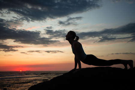 Silhouette of a woman doing yoga in dog muzzle-up pose on the twilight sea coast.