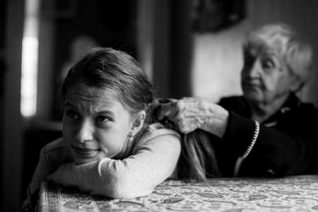 Grandmother braids her hair to a disgruntled little girl. Black and white picture.の写真素材