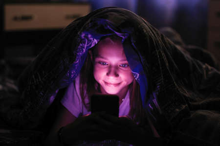Little cute girl at dark looks at smartphone under a blanket in bed.