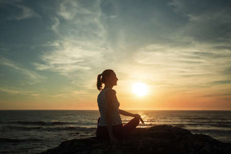 Silhouette woman practicing yoga on the sea beach at sunset.	の写真素材