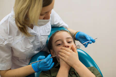 Female dentist doing teeth checkup of little girl looks worried with fear closes mouth with handssitting in a dental chair.  の写真素材