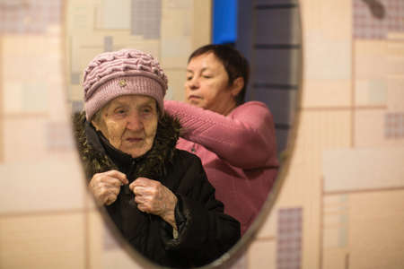Reflection in the mirror, a woman helps put on her old mother's coat.の写真素材