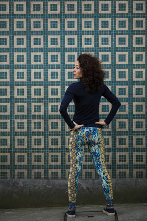 Portrait of a young Asian woman in leggings and sneakers on a city street, warm up near the wall....