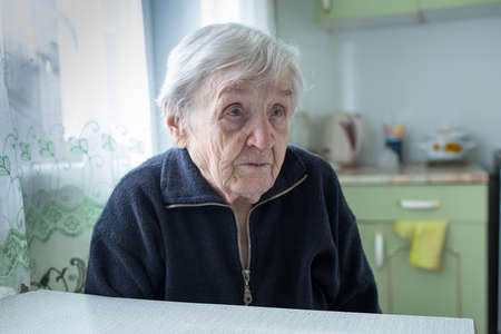Portrait of lonely old woman sitting in the kitchen by the window in her house.の写真素材