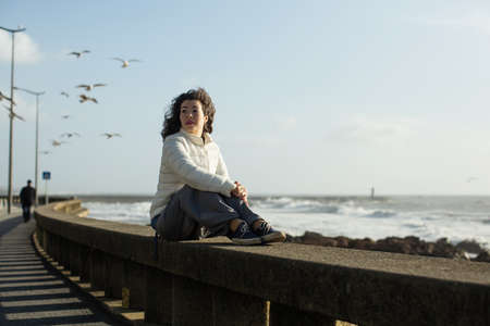 Multicultural woman sits on the waterfront during the surf and gulls around. Porto, Portugal.の写真素材