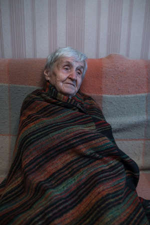 An old woman is sitting on the sofa wrapped in a blanket.の写真素材